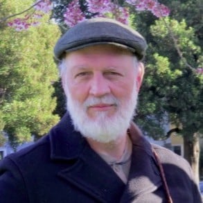 Dave Berry, an older man with a white beard and cap, stands outdoors in front of trees and blooming flowers, wearing a dark coat.