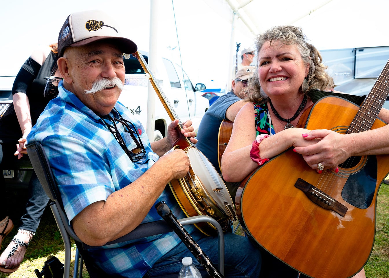 5B_CE_13444202_1740326953_6 Two smiling adults sit outdoors, one holding a banjo and the other an acoustic guitar, with other people and instruments visible in the background.