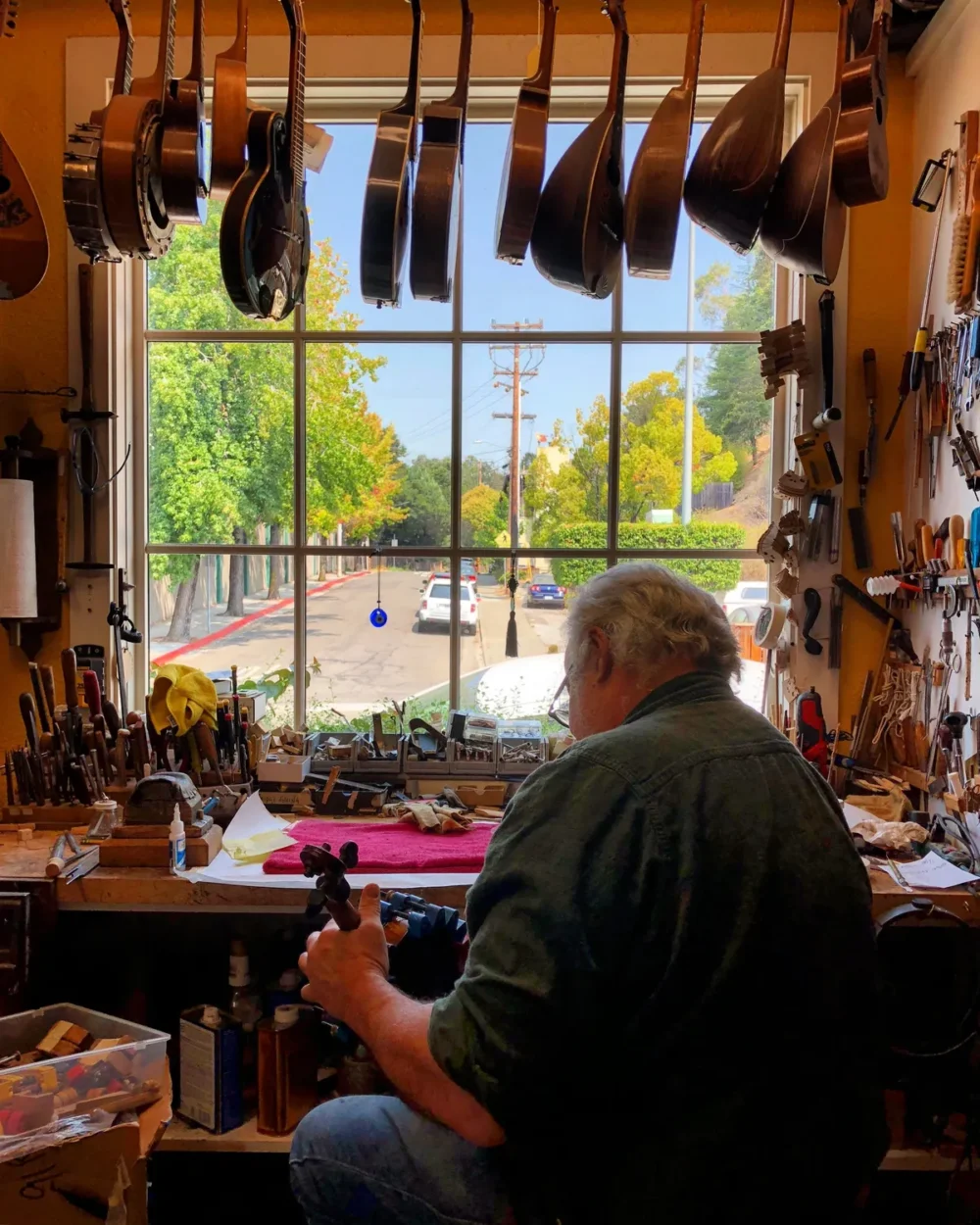 John Pedersen John Pedersen works on an instrument at a cluttered workbench in his workshop, with guitars hanging above and a window revealing a residential street and greenery outside.