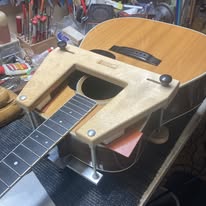 A guitar with its neck removed is on a workbench. A wooden clamp tool is attached to the body near the neck joint, surrounded by repair tools.