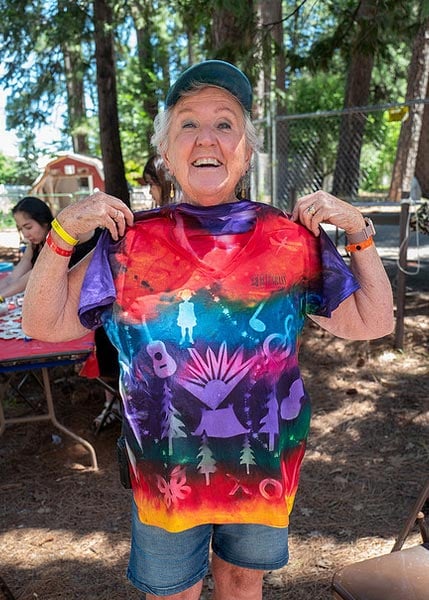 Kathleen-Rushing-SMC2026 Smiling older woman holding up a colorful, decorated t-shirt outdoors at what appears to be a craft event or fair.
