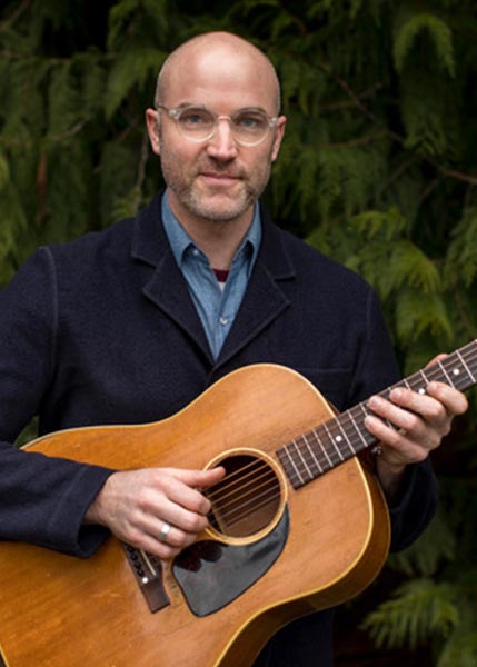 Eli-West-SMC2026 A man wearing glasses and a dark jacket stands outdoors, holding and playing an acoustic guitar with green foliage in the background.