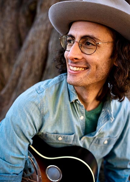 Mike-Witcher-SMC2026 Man wearing glasses and a wide-brimmed hat smiles while holding a guitar, sitting outdoors in front of a large tree.
