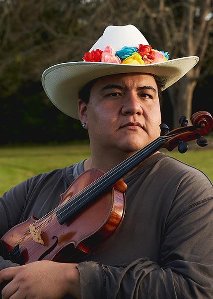 Nokosee-Fields-SMC2026 A person wearing a white hat decorated with colorful flowers holds a violin outdoors, with trees and grass in the background.