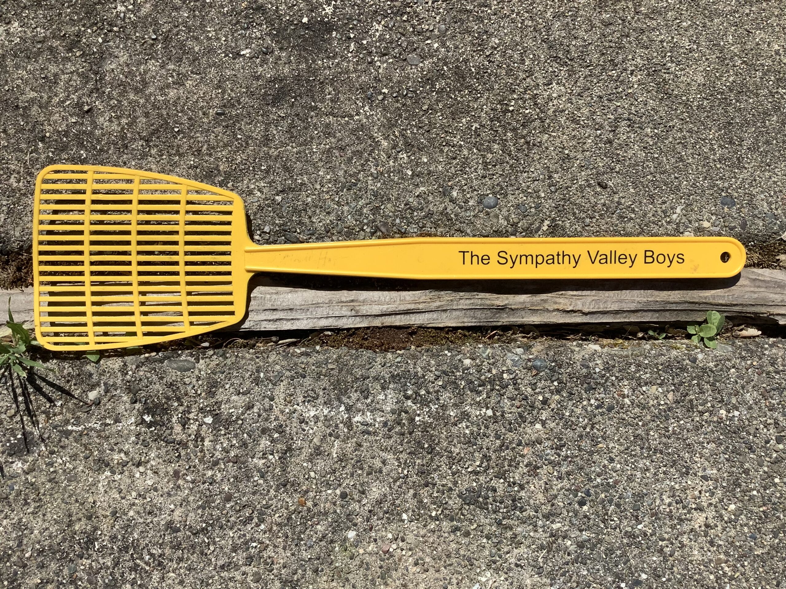 A yellow fly swatter with "Sympathy Valley Boys" printed on the handle, resting on a gray concrete surface beside a small wooden stick.