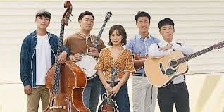 Five individuals, four men and one woman, are holding musical instruments including a double bass, banjo, mandolin, and guitars while standing in front of a light-colored wall in an Asian setting.