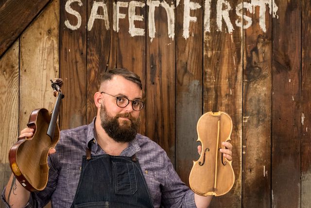 BGbio333small Brandon Godman A person with a beard and glasses holds a violin and a violin body in front of a wooden wall with the words "SAFETY FIRST" painted on it.