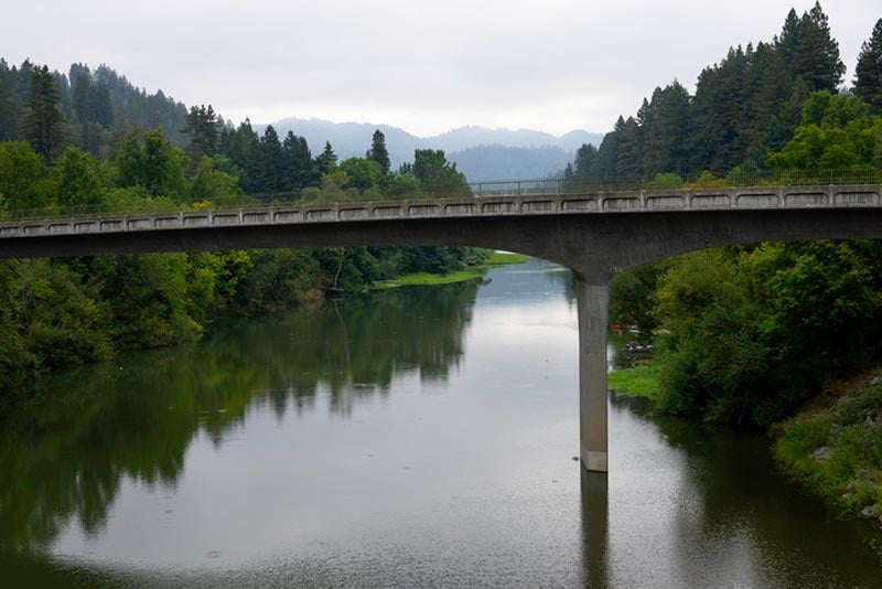 A concrete bridge spans a calm river surrounded by lush, forested hills under an overcast sky.
