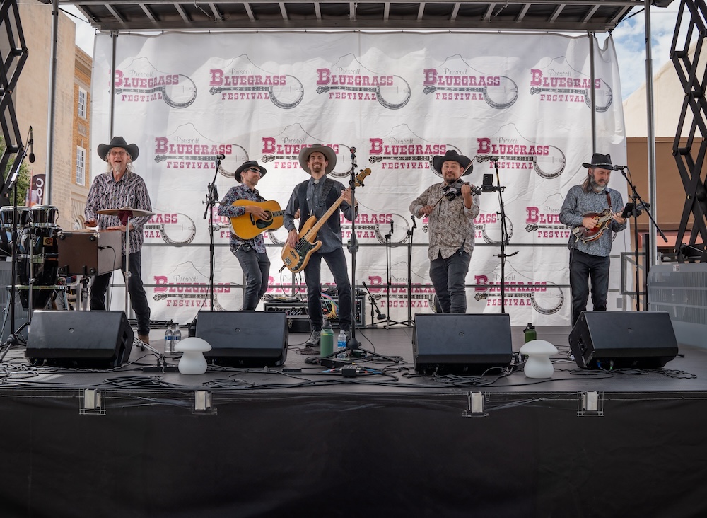 A five-member band performs on an outdoor stage at a bluegrass festival. Musicians play mandolin, guitar, bass, fiddle, and drums in front of a large festival banner.