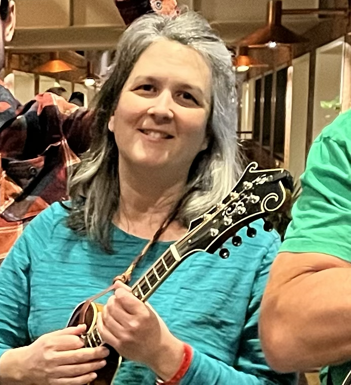 A person with gray hair is playing a mandolin, wearing a blue shirt and smiling.