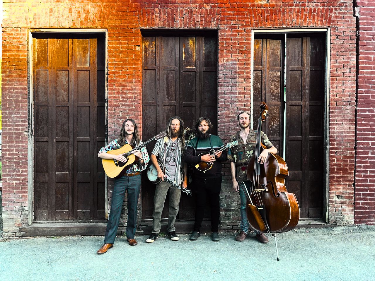 Four musicians stand against a brick wall, each holding an instrument: guitar, banjo, mandolin, and upright bass.