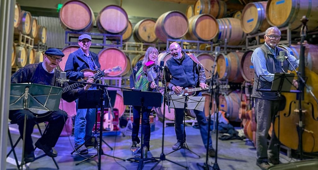 Five musicians perform in front of wine barrels, playing guitars, a banjo, a violin, and a stand-up bass. They are singing into microphones.