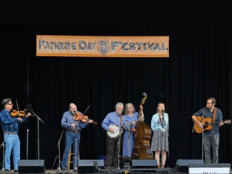 A bluegrass band performs on stage at the Father's Day Festival, featuring musicians with a banjo, guitars, fiddles, and a double bass.