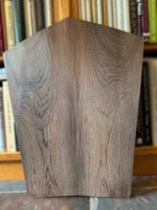 A Brazilian Rosewood object with distinct grain patterns is positioned upright in front of a blurred bookshelf filled with various books.