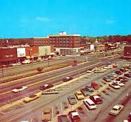 A parking lot filled with cars is in the foreground, while a wide road and several low-rise commercial buildings—once bustling with lint head workers—stretch into the background under a clear blue sky.