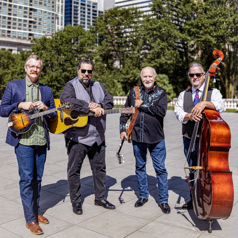 Four men stand outdoors holding musical instruments: mandolin, guitar, banjo, and upright bass, with city buildings and trees in the background.