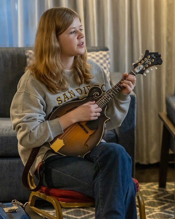 sophia sparks A person with long hair sits on a chair indoors, playing a mandolin and wearing a gray "San Diego" sweatshirt and jeans—one of the Young Pickers to Watch.