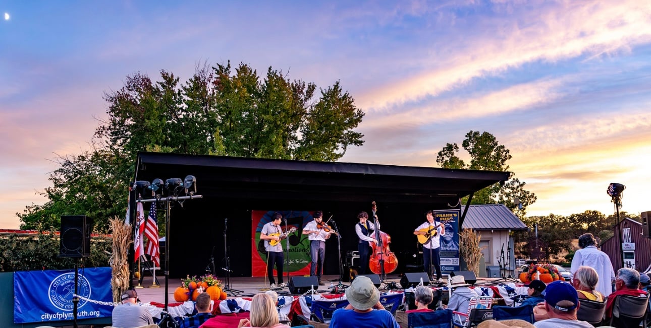 A band performs on an outdoor stage at sunset as an audience watches, with musical instruments and decorations visible.