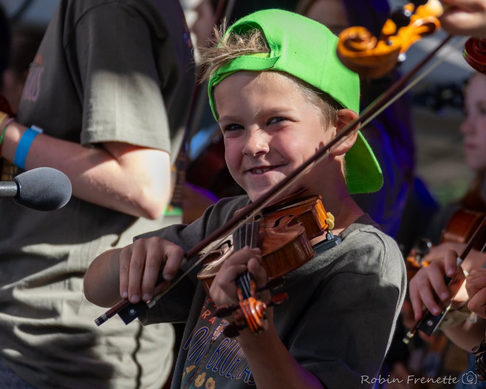 A young boy wearing a green cap plays the violin with a smile during a group outdoor performance.