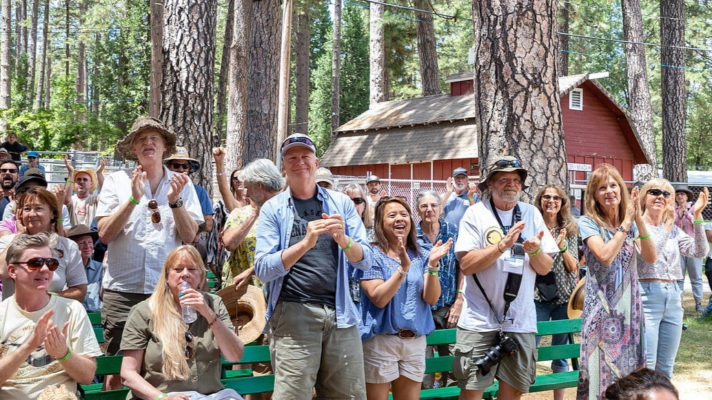 A group of people outdoors at a wooded venue clap and smile while standing and sitting on benches, with trees and a red building in the background.