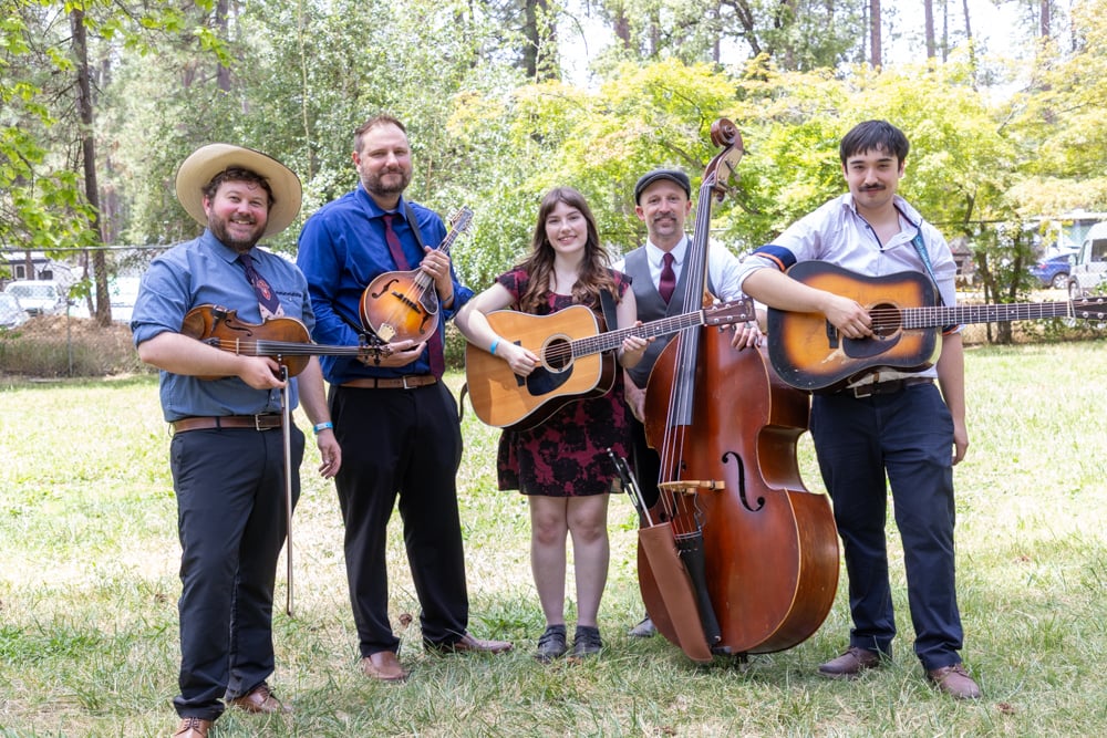 Five musicians stand outdoors with instruments including a fiddle, mandolin, guitar, upright bass, and another guitar, posing for a group photo in a grassy area with trees behind them.