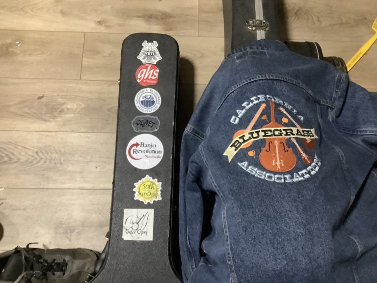 A guitar case with various music-related stickers sits next to a denim jacket featuring a California Bluegrass Association patch, evoking memories of summer vacation adventures, all resting on a wooden floor.