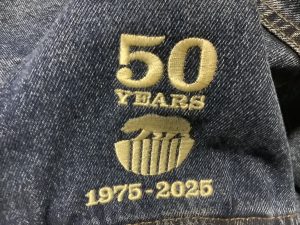 Close-up of denim fabric with embroidered text reading "50 Years," "1975-2025," and a logo featuring a hand and stylized lines—perfect for celebrating summer vacation milestones.