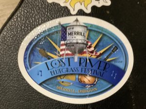 Oval sticker for Lost River Bluegrass Festival in Merrill, Oregon—perfect for summer vacation—features a water tower, American flag, guitar, banjo, and musical notes on a blue background.