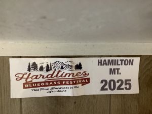 A sign reads "Hardtimes Bluegrass Festival: Old Time Bluegrass in the Mountains, Hamilton MT. 2025" with a mountain and trees graphic—an ideal summer vacation event for bluegrass lovers.