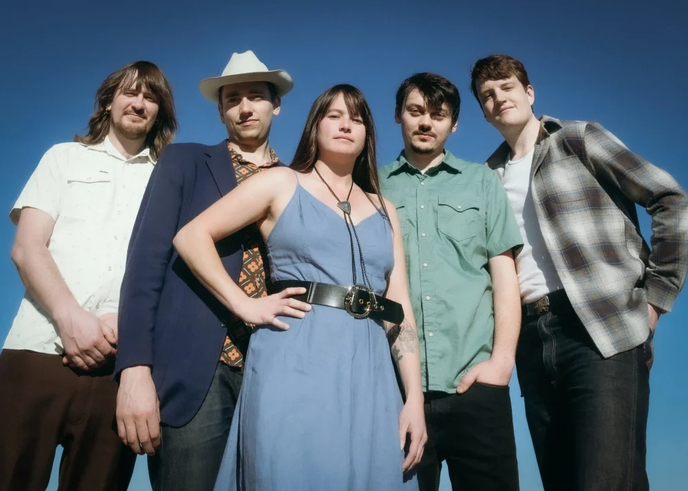 Five people stand outdoors against a clear blue sky, facing the camera. One person wears a hat, and another wears a dress; the rest are dressed in casual shirts and jackets.