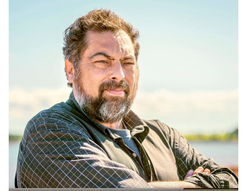 A man with a beard and mustache stands outdoors with arms crossed, wearing a checkered shirt and vest, with a blurred sky and water background.