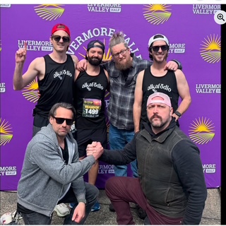 IMG_6695.jpeg Six men pose together in front of a purple Livermore Valley Half Marathon backdrop, with four wearing matching black "AlkalGold" tank tops. Two kneel and clasp hands in front.