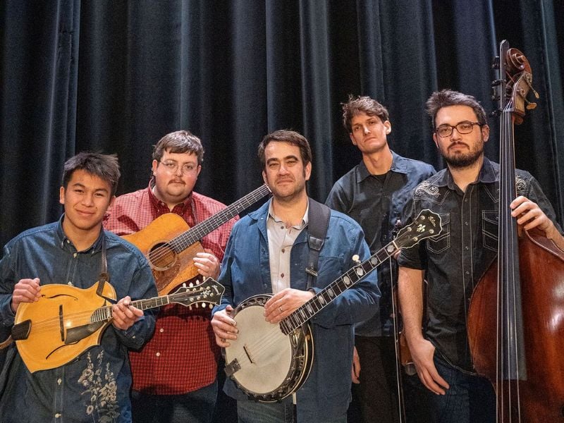 Five men stand in front of a dark curtain, each holding a musical instrument: a mandolin, acoustic guitar, banjo, fiddle, and upright bass.