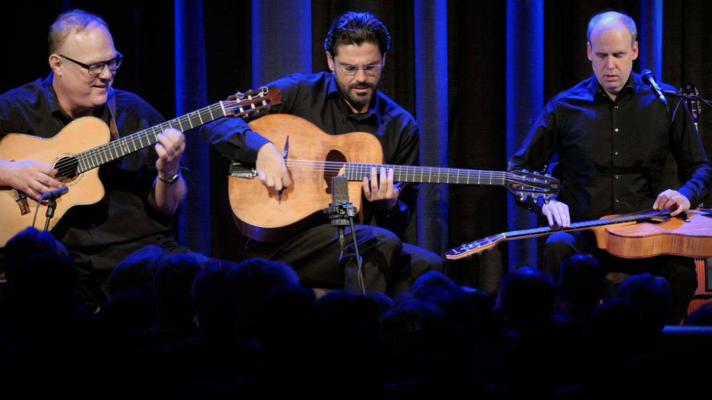 Three musicians in black clothing perform on stage with acoustic guitars and a lap steel guitar in front of a blue-lit curtain and seated audience.
