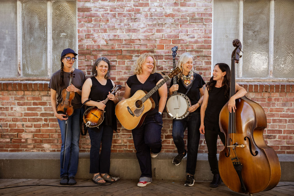 Five women stand in front of a brick wall, each holding a different musical instrument: fiddle, mandolin, guitar, banjo, and double bass. They are casually dressed and smiling.