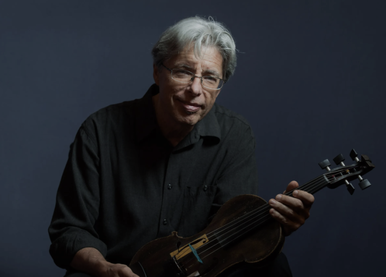 An older man with gray hair and glasses holds a violin while sitting against a dark background.