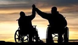Two people with disabilities in wheelchairs face each other and raise their arms for a high five against a sunset background.