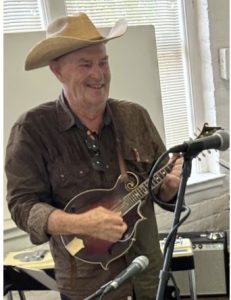 A man wearing a cowboy hat and brown shirt plays a mandolin in front of a microphone in a room with musical equipment and large windows.