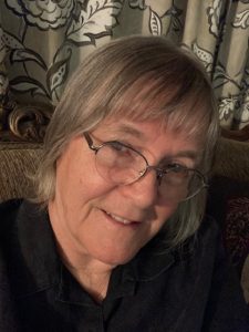 A person with gray hair and glasses smiles at the camera, seated on a couch with patterned curtains in the background.