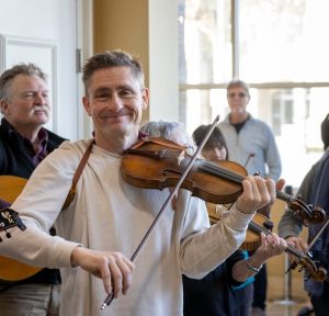A man in a white shirt plays the violin indoors, smiling at the camera, with several people and musicians visible in the background.