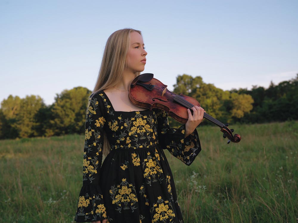Bronwyn-Keith-Hynes_web A woman in a black and yellow floral dress stands in a grassy field, holding a violin under her chin and looking to the side. Trees and blue sky are in the background.