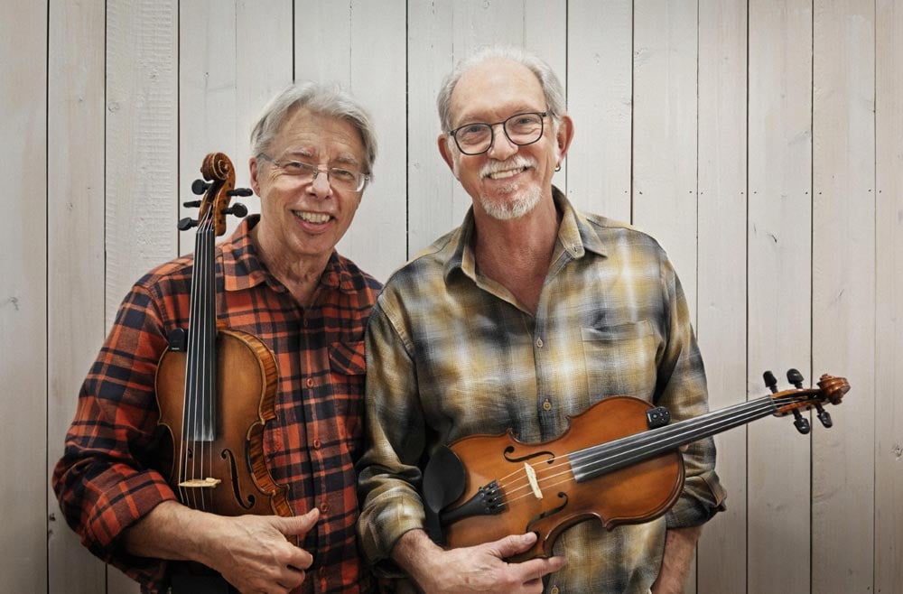 Bruce-Molsky-Darol-Anger_web Two older men in plaid shirts stand side by side, each holding a violin, and smiling in front of a light wooden wall.
