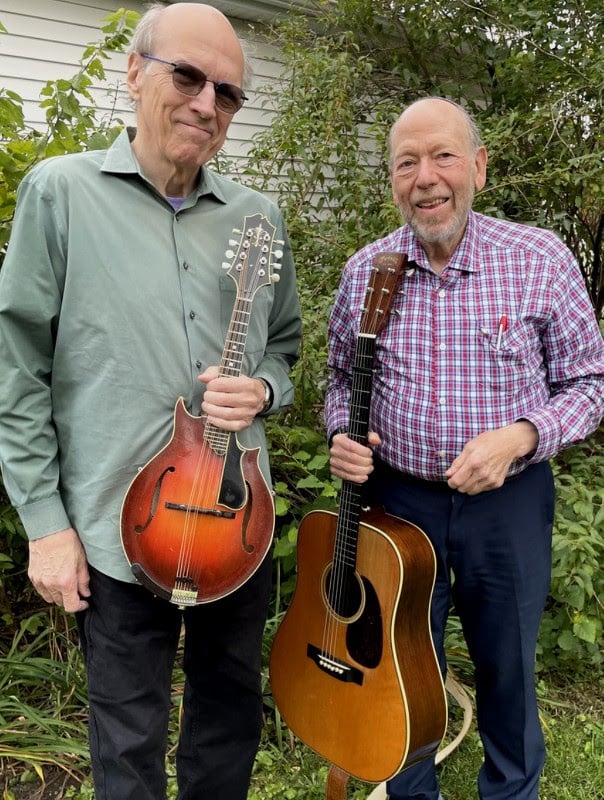 Two older men stand outdoors; one holds a mandolin, the other a guitar. They are in front of green bushes and a white building wall.