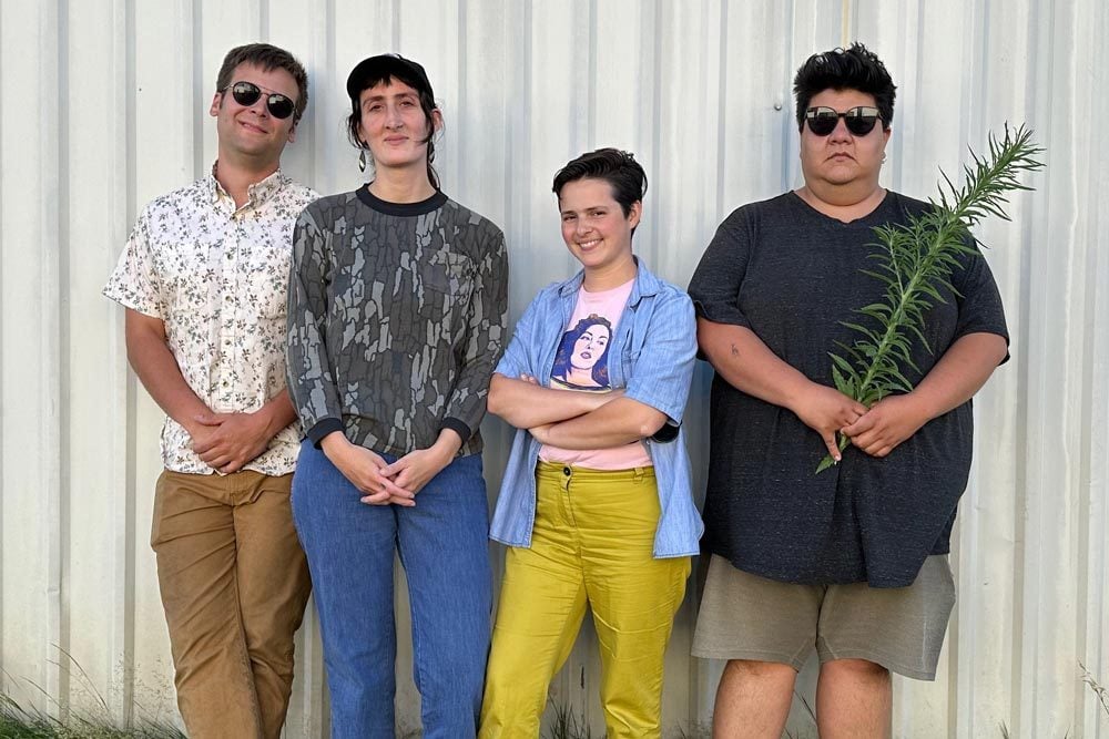 Hard-Drive_web Four people stand in a row outside against a metal wall; one holds a plant, and all have neutral or relaxed expressions.