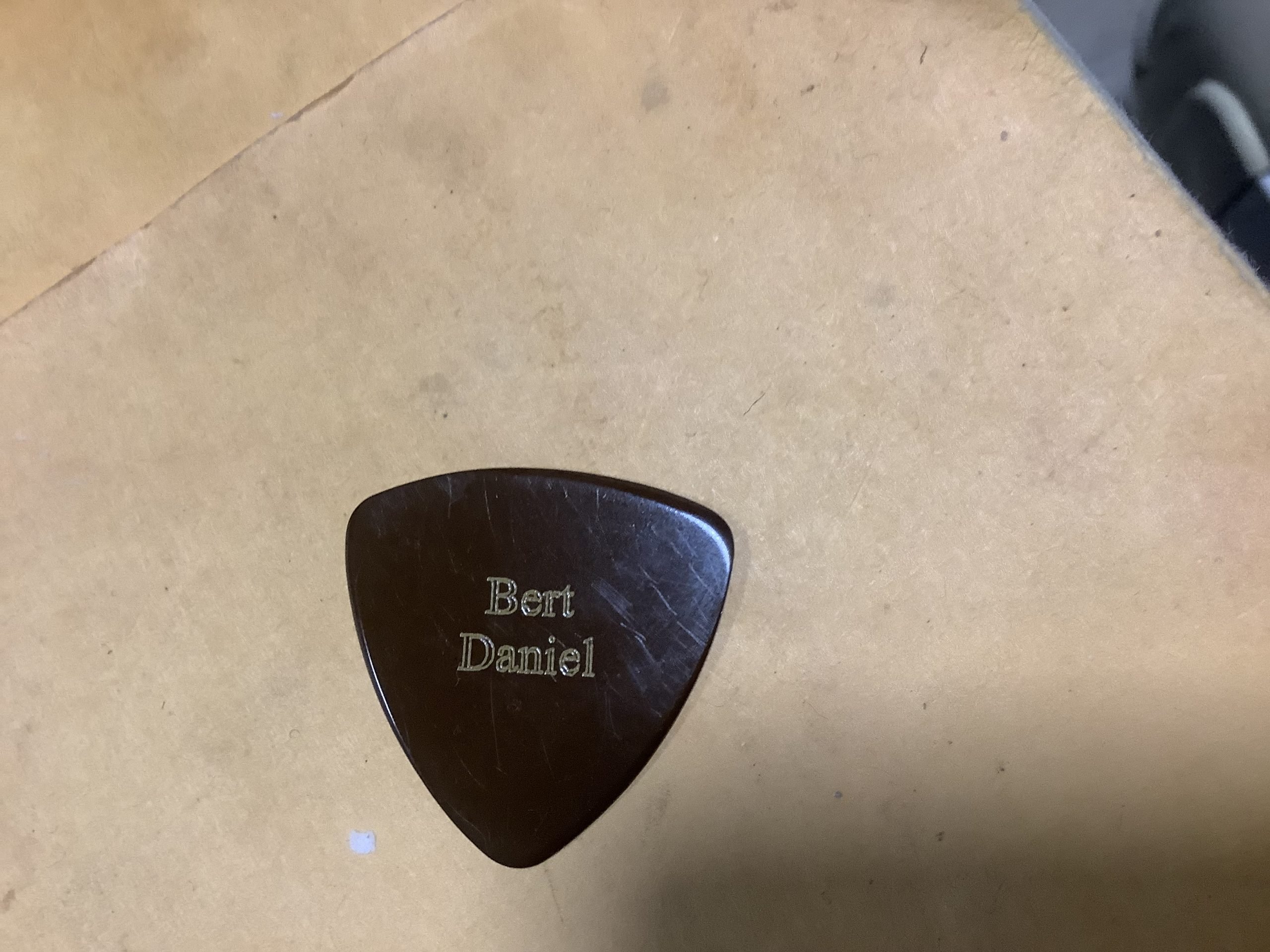A brown guitar pick engraved with the name "Bert Daniel" rests on a tan surface.