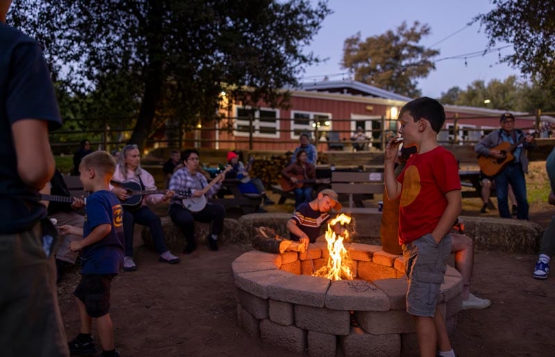 Children and adults sit around a campfire in the evening; some play guitars while others watch or stand nearby, with a building and trees in the background.
