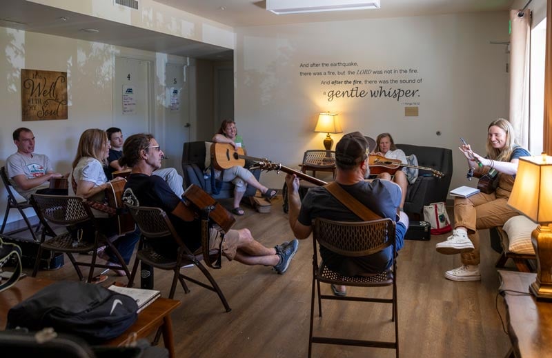 A group of people sits in a circle indoors playing guitars and singing, with a quote on the wall behind them and warm lighting in the room.