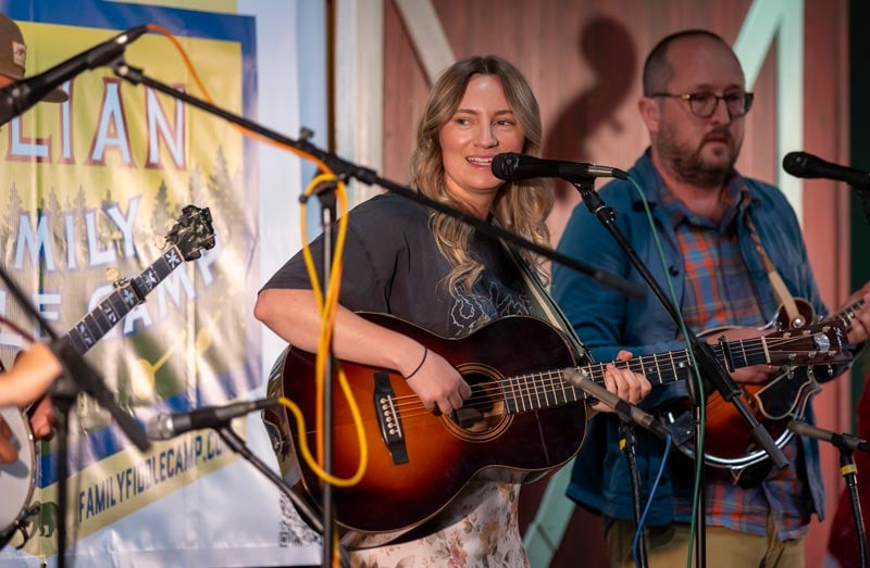 A woman plays an acoustic guitar and sings into a microphone on stage while a man beside her plays a mandolin.