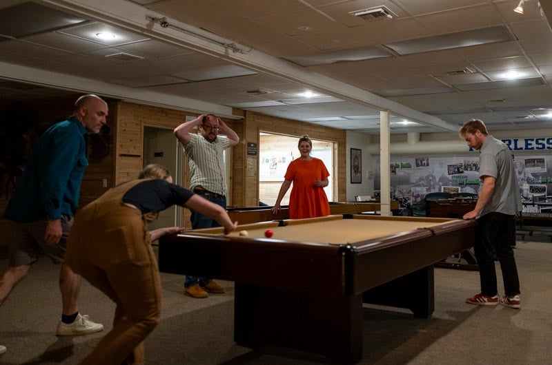 Five people are gathered around a pool table indoors, with one person taking a shot while others watch and react.