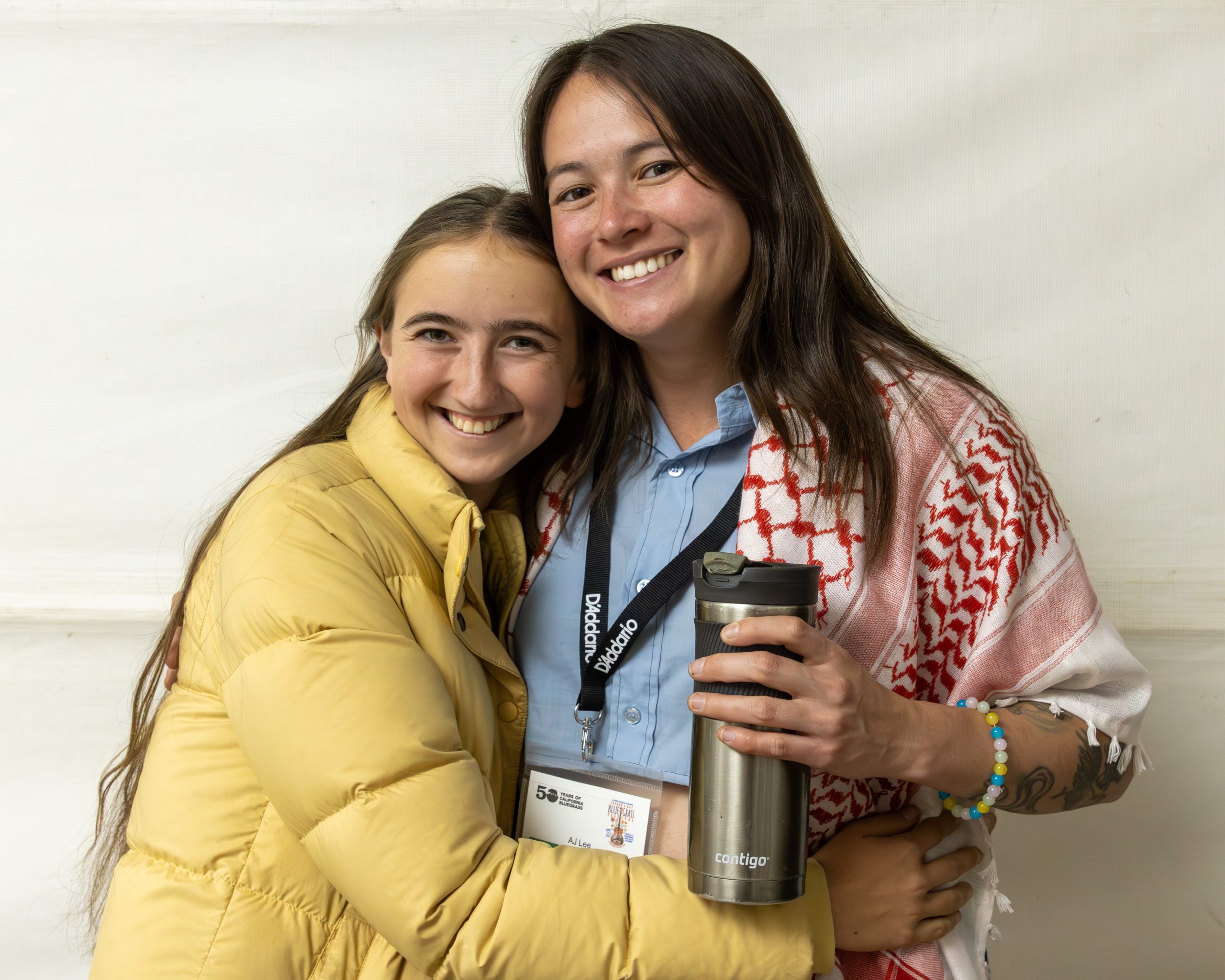 Two women standing side by side, smiling at the camera. One wears a yellow jacket, the other a red-patterned scarf and holds a travel mug. Both have visible name tags.